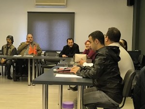 Fotoğrafta Antalya kent konseyinin toplantı odasında bir grup insan görülüyor. Masanın etrafında oturan kişiler, birbirleriyle konuşuyor veya dinliyor gibi görünüyor. Masanın üzerinde iki adet küçük Türk bayrağı yer alıyor. Arka planda, kapalı bir pencere ve üzerinde bir klima ünitesi bulunuyor. Katılımcılar arasında farklı yaş ve cinsiyetlerden insanlar var. Bazıları not alıyor gibi görünüyor. Ortam, resmi veya yarı resmi bir toplantı havasında.