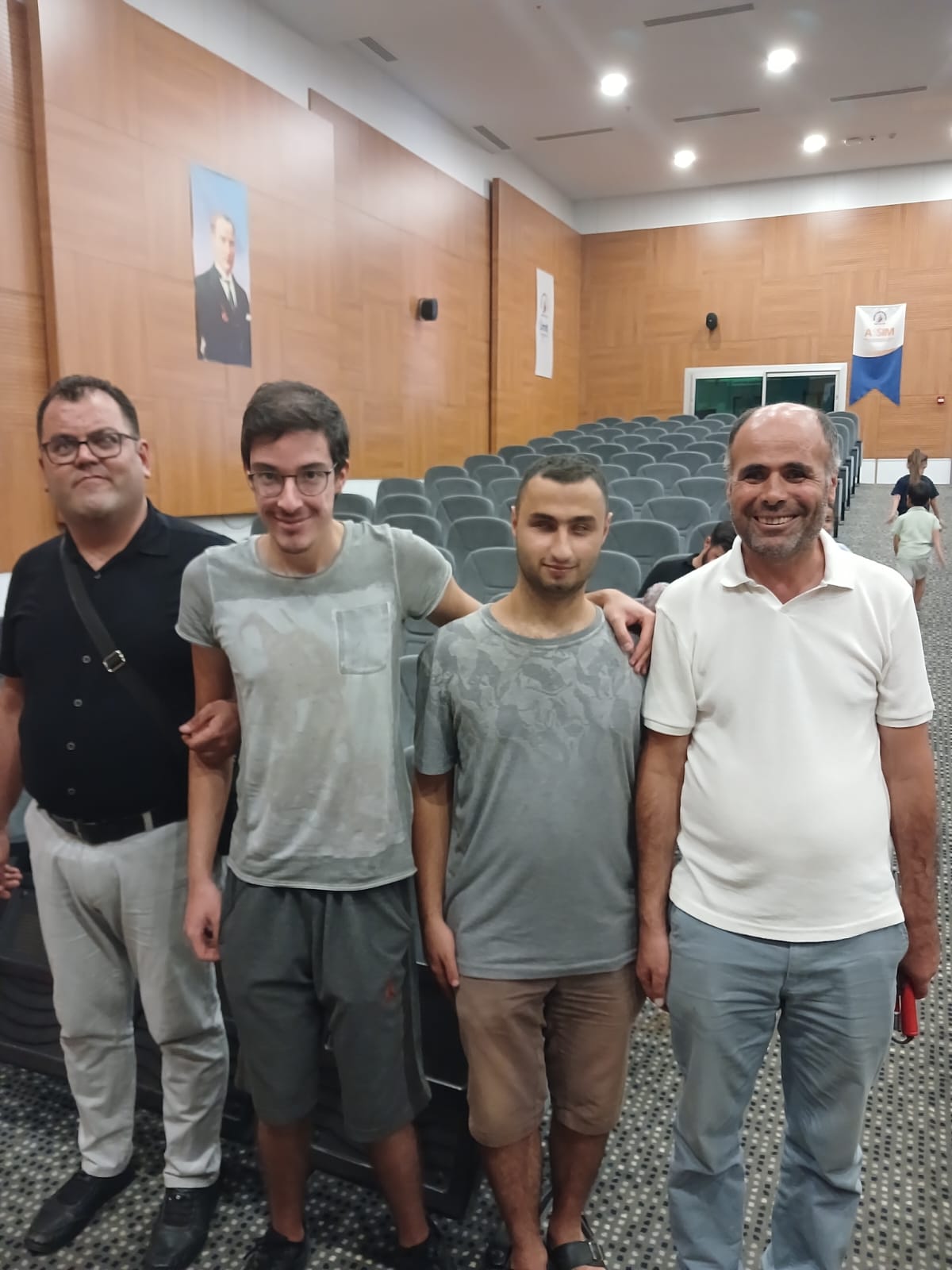 Fotoğrafta, bir arada poz veren Yönetim Kurulu üyeleri görülüyor. Sol başta duran Oğuz ÖZDEMİR, siyah bir tişört ve beyaz pantolon giymiş, gözlük takıyor ve omzunda bir çanta var. Yanındaki Ahmet Semih BİŞKİN, gri bir tişört ve gri şort giymiş, gözlük takıyor. Onun yanında duran Mert YAKIŞTIR, gri bir tişört ve kahverengi şort giymiş. En sağda ise Habib SİNCAR, beyaz bir tişört ve açık mavi pantolon giymiş, elinde kırmızı bir defter veya kitap tutuyor. Arka planda konferans salonunun detayları görülüyor, duvarlarda Atatürk'ün bir portresi ve bir afiş asılı. Oturma alanında gri koltuklar sıralanmış.