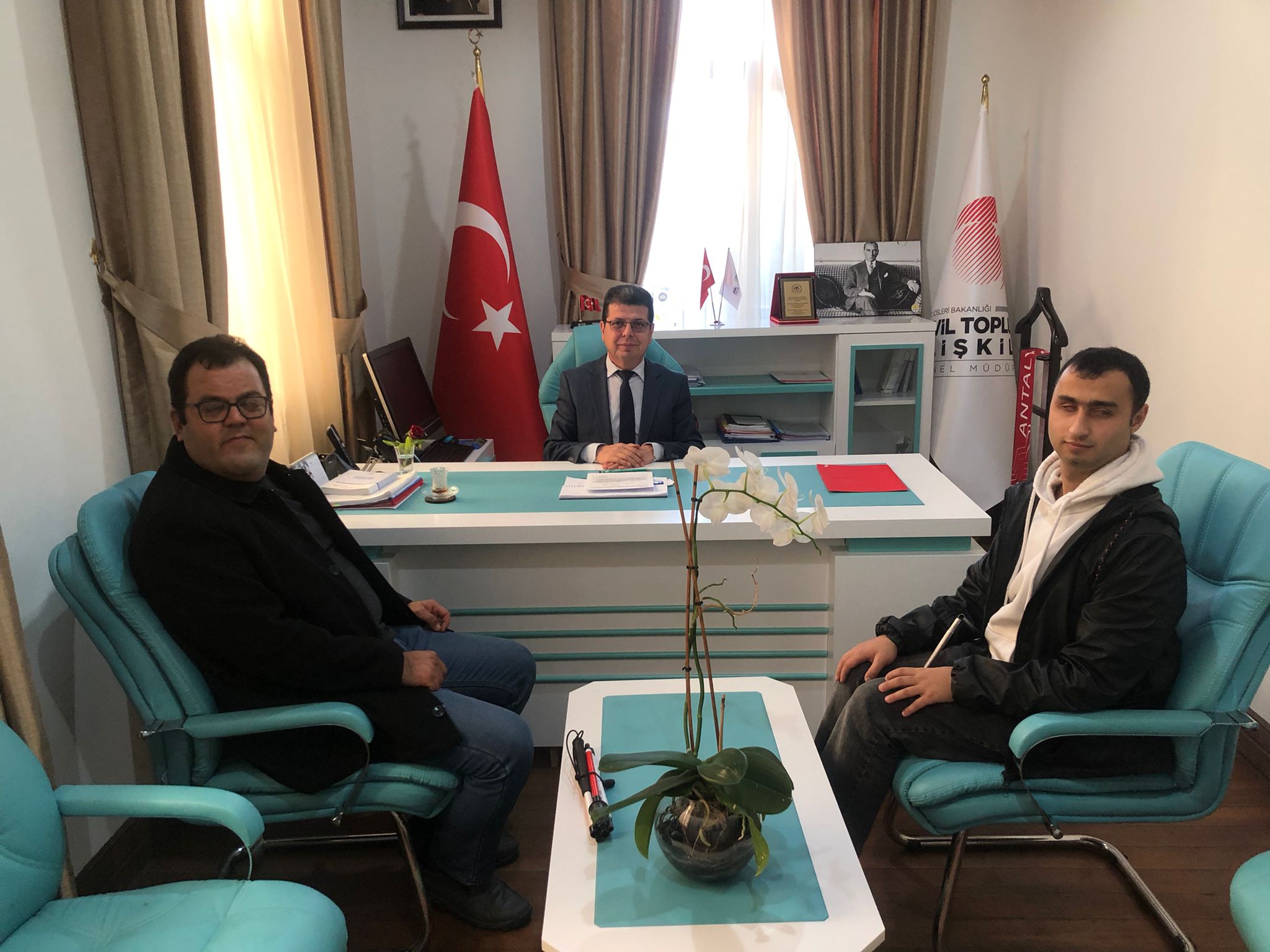 Fotoğrafta bir ofis ortamı görülüyor. Ortada bir masa var ve masanın arkasında takım elbise giymiş Hüseyin KURTAY oturuyor. Masanın üzerinde çeşitli belgeler ve bir bilgisayar bulunuyor. Masanın sol tarafında oturan kişi Oğuz ÖZDEMİR siyah bir ceket giymiş ve gözlük takıyor. Sağ tarafta oturan kişi ise Mert YAKIŞTIR beyaz bir kapüşonlu ve siyah bir ceket giymiş. Masanın üzerinde bir saksıda orkide çiçeği var. Arka planda Türk bayrağı ve Genel Müdürlüğün bayrağı da görülüyor. Ayrıca duvarda Atatürk'ün bir fotoğrafı asılı. Sağ arka planda ise "Sivil Toplumla İlişkiler Genel Müdürlüğü" yazılı bir pankart var.