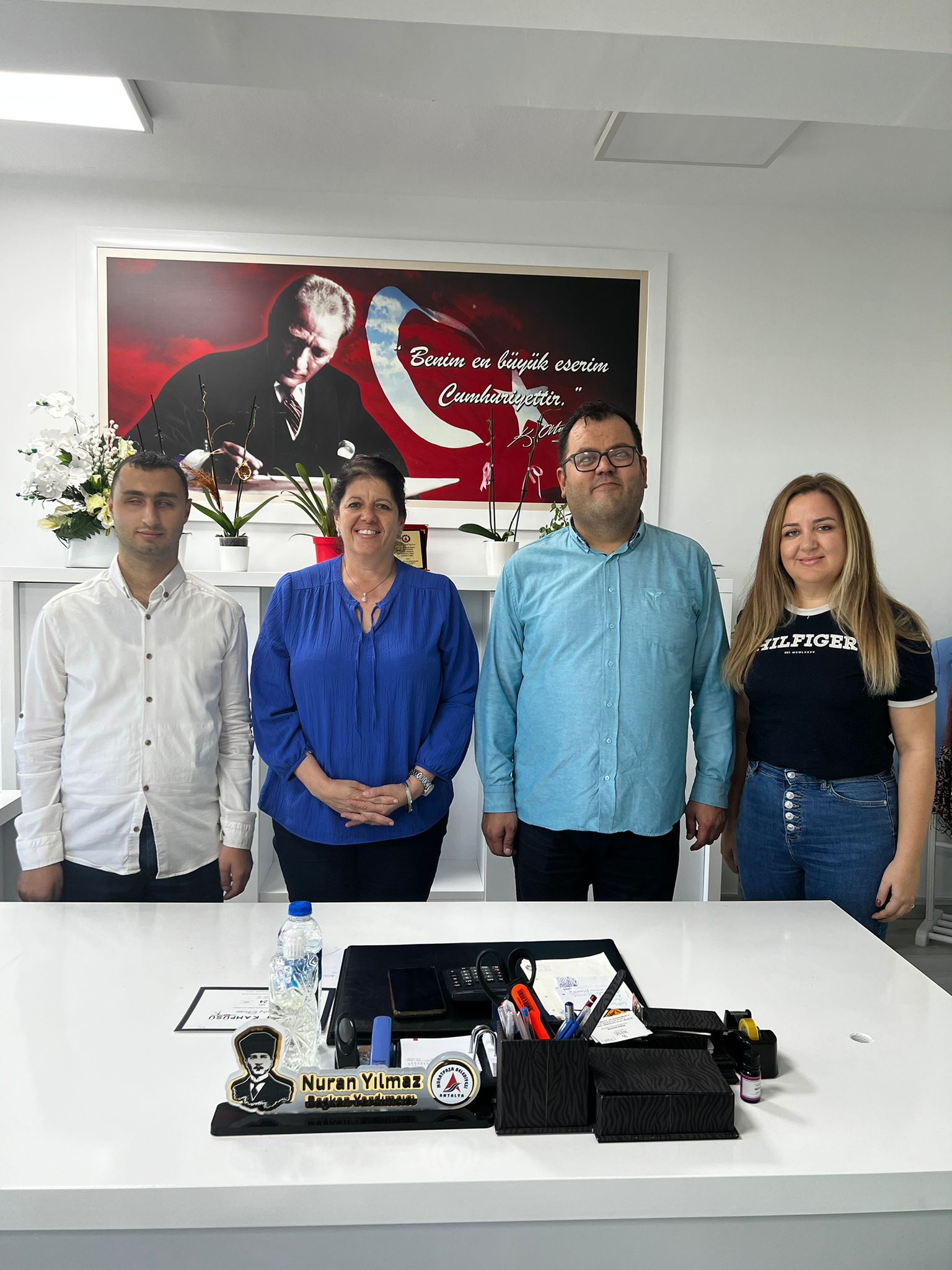 Fotoğrafta dört kişi bir masanın arkasında ayakta duruyor. Soldan sağa doğru, ilk kişi beyaz gömlek giymiş bir erkek. Yanında mavi bir bluz giymiş bir kadın var. Onun yanında açık mavi gömlek giymiş bir erkek duruyor. En sağda ise siyah tişört ve kot pantolon giymiş bir kadın var. Arkalarında, duvarda Atatürk'ün bir resmi ve "Benim en büyük eserim Cumhuriyettir." yazısı bulunuyor. Masanın üzerinde ise çeşitli ofis malzemeleri ve "Nuran Yılmaz" isminin yazılı olduğu bir isimlik var.
