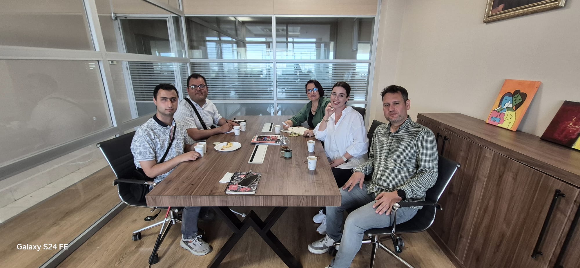 Masanın etrafındaki beş kişi, modern ofis sandalyelerinde oturuyor. Kişilerden üçü erkek, ikisi kadın. Herkes rahat bir şekilde oturuyor ve kameraya bakıyor. Soldan sağa doğru, ilk kişi beyaz desenli bir tişört giymiş ve çapraz bir çanta takmış. Yanındaki kişi gözlük takıyor ve beyaz bir gömlek giymiş. Ortadaki kadın yeşil bir ceket giymiş ve elinde bir kalem tutuyor. Yanındaki kadın beyaz bir gömlek giymiş ve gülümsüyor. En sağdaki erkek kareli bir gömlek giymiş ve elini masanın kenarına koymuş. Masanın üzerinde birkaç dergi, not defterleri, kahve fincanları ve beyaz kağıt bardaklar ile birkaç tabak içinde atıştırmalıklar bulunuyor. Arka plandaki cam bölmeler, ofis alanını ayırıyor ve dışarıdan gelen doğal ışık içeriye süzülüyor. Sağdaki dolabın üzerinde iki tablo var; biri soyut bir çift figürü, diğeri ise kırmızı tonlarda bir tasarım içeriyor.