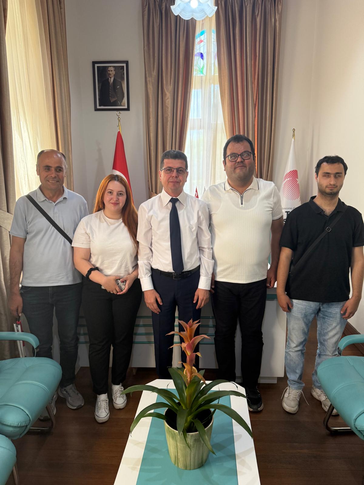 Beş kişilik bir grup, bir ofis ortamında ayakta duruyor. Ortadaki kişi takım elbise giymiş ve kravat takıyor, diğerleri ise daha rahat kıyafetler giymiş. Sol baştaki adamın omzunda bir çanta askısı var. Kadın, beyaz bir tişört ve siyah pantolon giymiş, elinde bir telefon tutuyor. Sağ baştaki iki adamdan biri beyaz bir tişört, diğeri ise siyah bir tişört giymiş. Arkada, duvarda Atatürk'ün bir portresi asılı ve Türk bayrağı ile başka bir bayrak yer alıyor. Ortada bir masa üzerinde saksıda bir bitki var. Oda, büyük pencerelerden gelen doğal ışıkla aydınlanıyor ve perdeler açık.